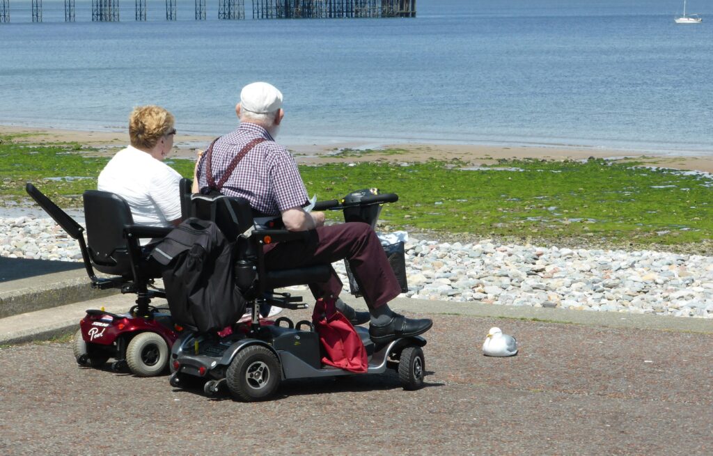An elderly couple enjoys a seaside view while seated on mobility scooters, emphasizing leisure and accessibility.