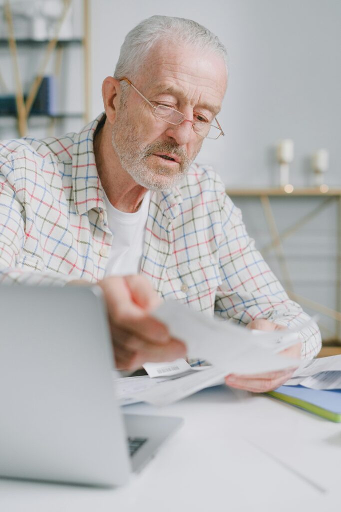 Elderly man with glasses reading papers at a table, using a laptop indoors.