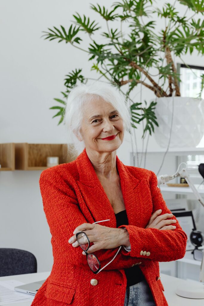 Elderly woman in red blazer standing confidently in modern office setting, holding eyeglasses.